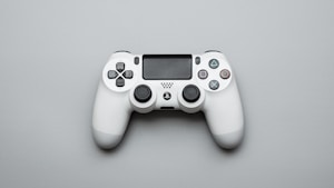 A white gaming controller is positioned on a light gray surface. The controller features dual analog sticks, a directional pad, and iconic four-button layout with distinct symbols. The design is sleek and modern, with additional features such as triggers and a central touchpad.