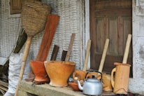 A collection of traditional wooden kitchen utensils and tools is arranged in front of a rustic wooden door. The assortment includes wooden mortars, pestles, a silver kettle, and a large woven basket. The background features textured, weathered walls adding to the rustic charm.