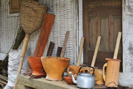 A collection of traditional wooden kitchen utensils and tools is arranged in front of a rustic wooden door. The assortment includes wooden mortars, pestles, a silver kettle, and a large woven basket. The background features textured, weathered walls adding to the rustic charm.