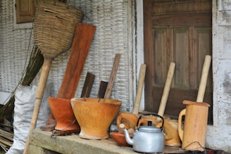 A collection of traditional wooden kitchen utensils and tools is arranged in front of a rustic wooden door. The assortment includes wooden mortars, pestles, a silver kettle, and a large woven basket. The background features textured, weathered walls adding to the rustic charm.