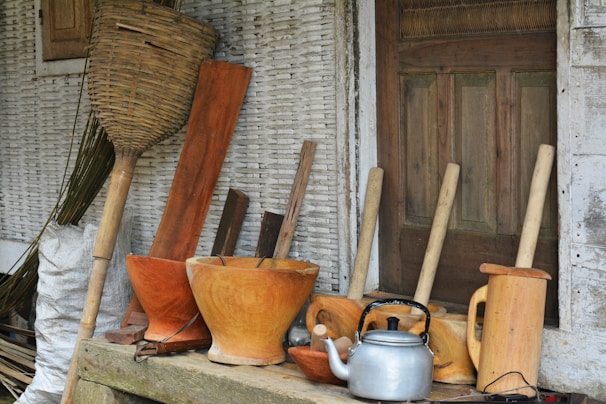 A collection of traditional wooden kitchen utensils and tools is arranged in front of a rustic wooden door. The assortment includes wooden mortars, pestles, a silver kettle, and a large woven basket. The background features textured, weathered walls adding to the rustic charm.
