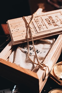 A wooden box partially opened to reveal a textured fabric with printed text. A gold chain is elegantly draped over the box, creating a stylish and sophisticated ambiance. Next to the box, a golden watch glimmers under the warm lighting, adding a hint of luxury and timelessness.
