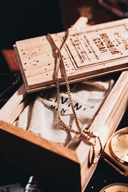 A wooden box partially opened to reveal a textured fabric with printed text. A gold chain is elegantly draped over the box, creating a stylish and sophisticated ambiance. Next to the box, a golden watch glimmers under the warm lighting, adding a hint of luxury and timelessness.