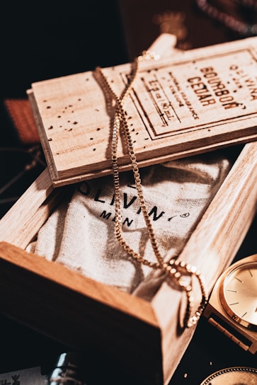 A wooden box partially opened to reveal a textured fabric with printed text. A gold chain is elegantly draped over the box, creating a stylish and sophisticated ambiance. Next to the box, a golden watch glimmers under the warm lighting, adding a hint of luxury and timelessness.