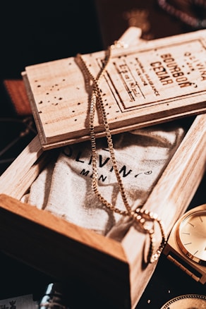 A wooden box partially opened to reveal a textured fabric with printed text. A gold chain is elegantly draped over the box, creating a stylish and sophisticated ambiance. Next to the box, a golden watch glimmers under the warm lighting, adding a hint of luxury and timelessness.
