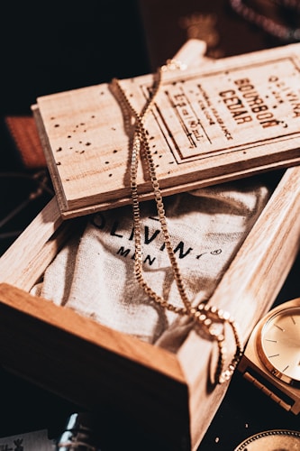 A wooden box partially opened to reveal a textured fabric with printed text. A gold chain is elegantly draped over the box, creating a stylish and sophisticated ambiance. Next to the box, a golden watch glimmers under the warm lighting, adding a hint of luxury and timelessness.