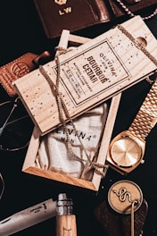 An arrangement of men's accessories including a wooden box labeled 'Bourbon Cedar' with a chain draped over it. Surrounding items include a gold wristwatch, a leather wallet, sunglasses, and a knife with a wooden handle. The composition suggests a theme of luxury and sophistication.