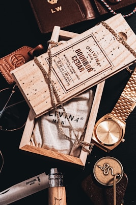 An arrangement of men's accessories including a wooden box labeled 'Bourbon Cedar' with a chain draped over it. Surrounding items include a gold wristwatch, a leather wallet, sunglasses, and a knife with a wooden handle. The composition suggests a theme of luxury and sophistication.