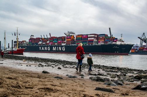 A large container ship named Yang Ming is docked near a port, loaded with multicolored shipping containers. In the foreground, a woman in a red coat and a child in a blue hat walk along a rocky beach. Other ships and cranes can be seen in the background under a cloudy sky.