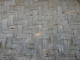 A close-up view of a woven bamboo texture, featuring a pattern of interlocking rectangular strips arranged in a criss-cross design. The bamboo is aged, displaying a faded, light grey color with slight discoloration and natural imperfections.