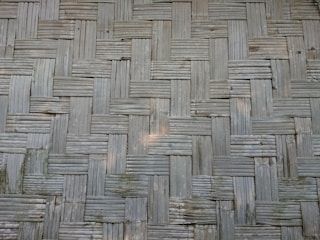 A close-up view of a woven bamboo texture, featuring a pattern of interlocking rectangular strips arranged in a criss-cross design. The bamboo is aged, displaying a faded, light grey color with slight discoloration and natural imperfections.