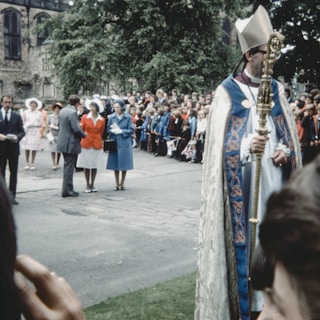 A large outdoor gathering with a diverse crowd lined up along the street. In the foreground, a figure dressed in ceremonial robes and a mitre holds an ornate staff. The crowd in the background appears to be watching a formal event, with some people holding flags. The attire suggests a formal occasion, possibly involving a religious or state ceremony, with people dressed in suits, hats, and coats.