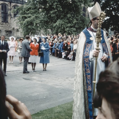 A large outdoor gathering with a diverse crowd lined up along the street. In the foreground, a figure dressed in ceremonial robes and a mitre holds an ornate staff. The crowd in the background appears to be watching a formal event, with some people holding flags. The attire suggests a formal occasion, possibly involving a religious or state ceremony, with people dressed in suits, hats, and coats.