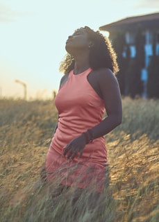 A serene woman in a soft pink dress standing barefoot in a field of tall grass under a pastel sunset sky.
