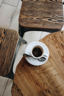 A cup of coffee sits on a wooden table with intricate grain patterns. Two chairs with similar wooden designs are nearby, creating a cohesive and rustic aesthetic.