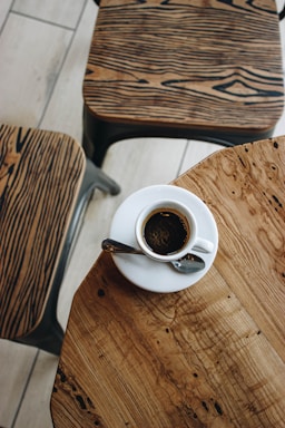 A cup of coffee sits on a wooden table with intricate grain patterns. Two chairs with similar wooden designs are nearby, creating a cohesive and rustic aesthetic.