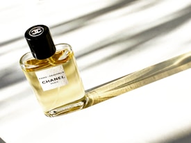 A bottle of Chanel perfume with a black cap and a label reading 'Paris - Deauville Chanel Paris.' It casts a long shadow on a surface with soft stripes of light creating an elegant and minimalist composition.