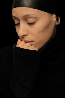 A person with closed eyes wearing a black headscarf and a black long-sleeved shirt rests their hand near their mouth, appearing thoughtful. The person has earphones in their ear.