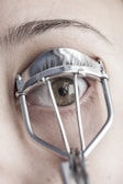 A close-up view of an eye with an eyelash curler applied. The eyelashes are partially visible through the metal frame of the curler, and the skin and eyebrow are in focus.