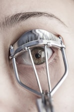 A close-up view of an eye with an eyelash curler applied. The eyelashes are partially visible through the metal frame of the curler, and the skin and eyebrow are in focus.