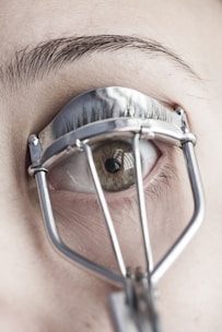 A close-up view of an eye with an eyelash curler applied. The eyelashes are partially visible through the metal frame of the curler, and the skin and eyebrow are in focus.