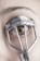 A close-up view of an eye with an eyelash curler applied. The eyelashes are partially visible through the metal frame of the curler, and the skin and eyebrow are in focus.