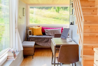 A cozy and well-lit small living space with a wooden staircase leading upwards. The room features a daybed adorned with colorful pillows in yellow and pink hues. A small desk with a chair is positioned near the bed, accompanied by a laptop, tablet, and other small items. Large windows provide a view of a lush, green outdoor landscape, allowing plenty of natural light to fill the space. Books are stacked near the windowsill, adding to the homey atmosphere.