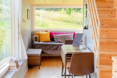 A cozy and well-lit small living space with a wooden staircase leading upwards. The room features a daybed adorned with colorful pillows in yellow and pink hues. A small desk with a chair is positioned near the bed, accompanied by a laptop, tablet, and other small items. Large windows provide a view of a lush, green outdoor landscape, allowing plenty of natural light to fill the space. Books are stacked near the windowsill, adding to the homey atmosphere.