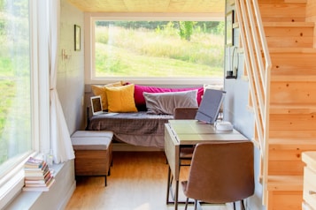 A cozy and well-lit small living space with a wooden staircase leading upwards. The room features a daybed adorned with colorful pillows in yellow and pink hues. A small desk with a chair is positioned near the bed, accompanied by a laptop, tablet, and other small items. Large windows provide a view of a lush, green outdoor landscape, allowing plenty of natural light to fill the space. Books are stacked near the windowsill, adding to the homey atmosphere.