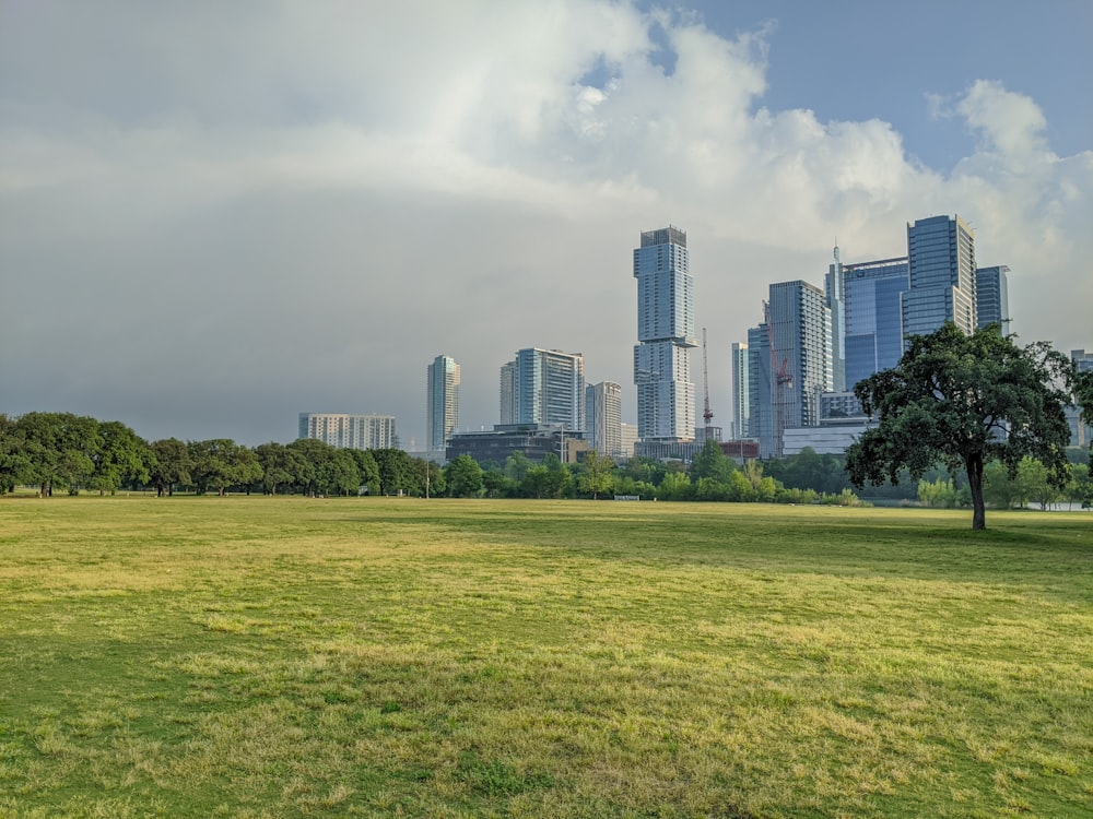 green grass field near city buildings under white clouds during daytime