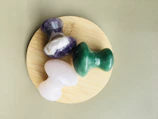 Three polished crystal stones, one purple and white, one green, and one pink, are arranged on a round, light wooden surface. The background is a textured, pale green material.