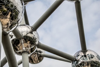 Geometric metal spheres are interconnected by large cylindrical beams against a cloudy sky background. The mirrored surfaces of the spheres reflect light and obscure shapes, creating a futuristic architectural design.