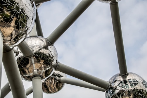 Geometric metal spheres are interconnected by large cylindrical beams against a cloudy sky background. The mirrored surfaces of the spheres reflect light and obscure shapes, creating a futuristic architectural design.