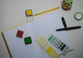 An open notebook with a yellow border is placed on a desk, accompanied by a yellow ink pad and a green ink pad. A pen is lying next to the notebook, and some adhesive decorative stickers in green and yellow are also present. A small succulent plant in a terracotta pot and a roll of green washi tape are situated nearby, while a patterned pencil rests close to the ink pads.