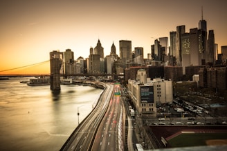 A panoramic view of a cityscape featuring a skyline with numerous skyscrapers. The Brooklyn Bridge is prominently visible, stretching across a calm river. The scene is captured during sunset, with a warm glow illuminating the buildings. Light trails from vehicles suggest a busy highway along the waterfront.