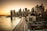 A panoramic view of a cityscape featuring a skyline with numerous skyscrapers. The Brooklyn Bridge is prominently visible, stretching across a calm river. The scene is captured during sunset, with a warm glow illuminating the buildings. Light trails from vehicles suggest a busy highway along the waterfront.
