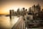 A panoramic view of a cityscape featuring a skyline with numerous skyscrapers. The Brooklyn Bridge is prominently visible, stretching across a calm river. The scene is captured during sunset, with a warm glow illuminating the buildings. Light trails from vehicles suggest a busy highway along the waterfront.