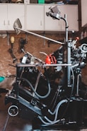 A complex, industrial machine with various levers, hoses, and mechanical components is situated in a workshop. The background features a pegboard wall with hanging tools and overhead cabinets for storage. A red oil can is placed on a flat surface of the machine.