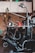 A complex, industrial machine with various levers, hoses, and mechanical components is situated in a workshop. The background features a pegboard wall with hanging tools and overhead cabinets for storage. A red oil can is placed on a flat surface of the machine.