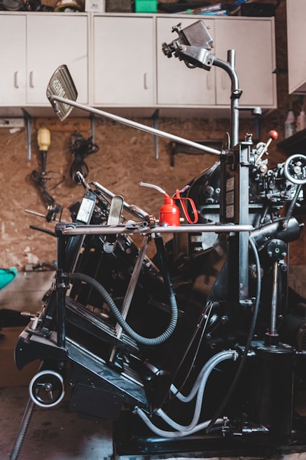 A complex, industrial machine with various levers, hoses, and mechanical components is situated in a workshop. The background features a pegboard wall with hanging tools and overhead cabinets for storage. A red oil can is placed on a flat surface of the machine.