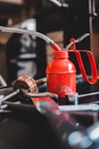 A red oil can with a metal spout and handle is surrounded by various tools, including a wrench and what appears to be a piece of metal with holes. The background is blurred, focusing attention on the oil can and tools, suggesting a workshop or garage setting.