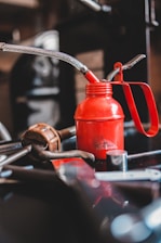 A red oil can with a metal spout and handle is surrounded by various tools, including a wrench and what appears to be a piece of metal with holes. The background is blurred, focusing attention on the oil can and tools, suggesting a workshop or garage setting.