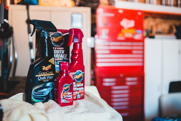Several car care products, including bottles of wax and detailing spray, are placed on a soft cloth in a garage setting. A red tool chest stands in the background, slightly out of focus, against a backdrop of shelves and storage areas. The products feature prominent labels and branding.