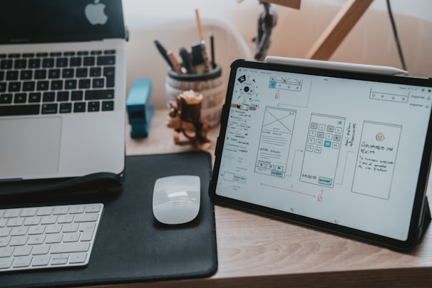 A workspace featuring a wooden desk with a laptop partially visible on the left and a digital tablet displaying wireframe designs on the right. There is a white computer mouse on a black mousepad next to a white keyboard. In the background, some stationery items like pens and pencils are held in a container, and a small wooden figurine is situated beside them.