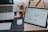 A workspace featuring a wooden desk with a laptop partially visible on the left and a digital tablet displaying wireframe designs on the right. There is a white computer mouse on a black mousepad next to a white keyboard. In the background, some stationery items like pens and pencils are held in a container, and a small wooden figurine is situated beside them.