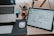 A workspace featuring a wooden desk with a laptop partially visible on the left and a digital tablet displaying wireframe designs on the right. There is a white computer mouse on a black mousepad next to a white keyboard. In the background, some stationery items like pens and pencils are held in a container, and a small wooden figurine is situated beside them.