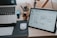A workspace featuring a wooden desk with a laptop partially visible on the left and a digital tablet displaying wireframe designs on the right. There is a white computer mouse on a black mousepad next to a white keyboard. In the background, some stationery items like pens and pencils are held in a container, and a small wooden figurine is situated beside them.
