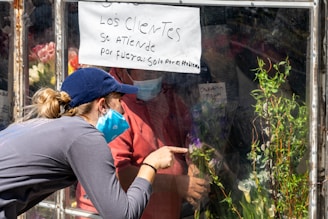 A person in a navy cap and mask points through a glass window at someone inside wearing a mask, surrounded by plants and flowers. A handwritten sign on the window in Spanish addresses customers about service changes, likely due to an issue.