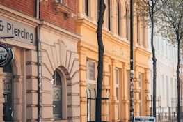 An urban street scene features a row of classic European-style buildings with ornate architectural details and large arched windows. The facade of a tattoo and piercing shop is prominent in the foreground. The buildings are painted in warm shades of beige, yellow, and orange. Leafy trees line the street, and iron fences are visible along the sidewalks.