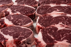 Several raw steak cuts of red meat with marbling are arranged in a neat, overlapping pattern. The meat appears fresh and ready for cooking.